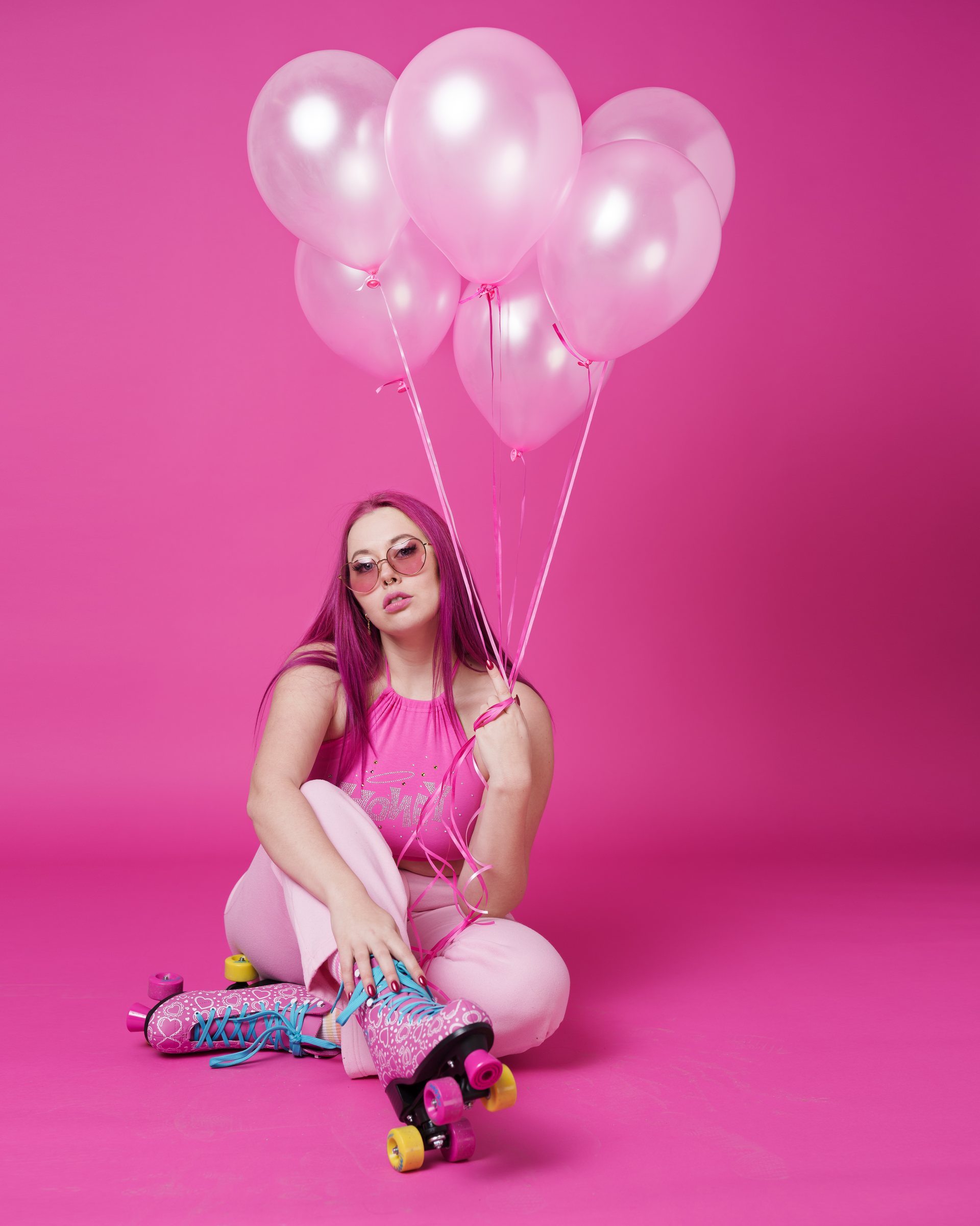 Frau mit pinkem Haar und Rollschuhen sitzt vor pinkem Hintergrund mit Luftballons